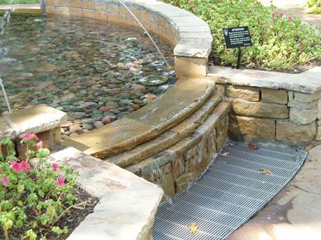 The gap in the side of the flower bed drains the water in the fountain, several leaves on the steel grating.